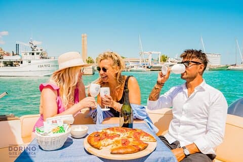 Three friends enjoy an aperitif on a boat in the sun. A man in sunglasses and white shirt sips a cocktail, while a woman with long hair and black top giggles, they are happy.